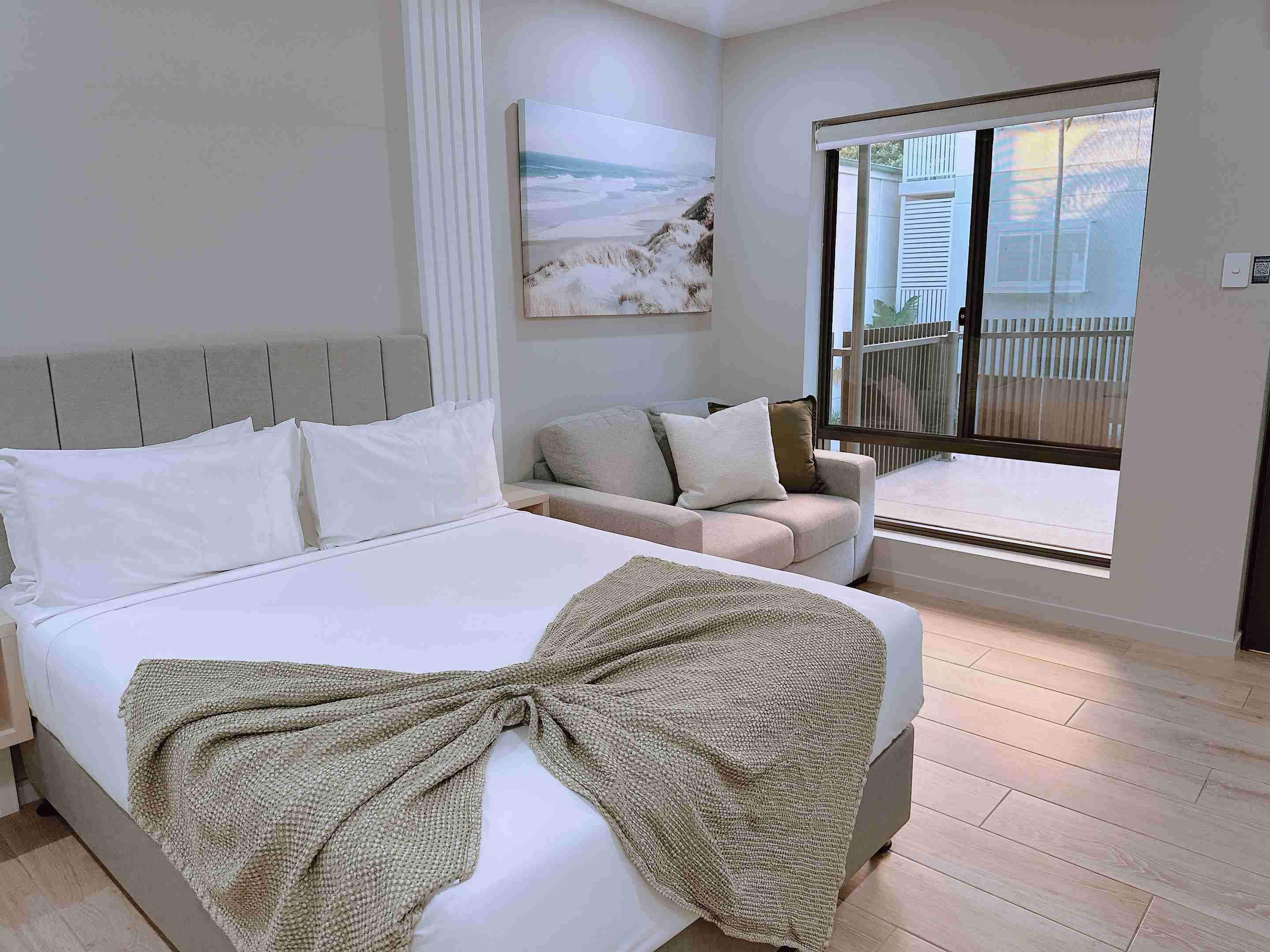 A bedroom with a bed, a sofa, a window with a view of the outside, and a painting on the wall.