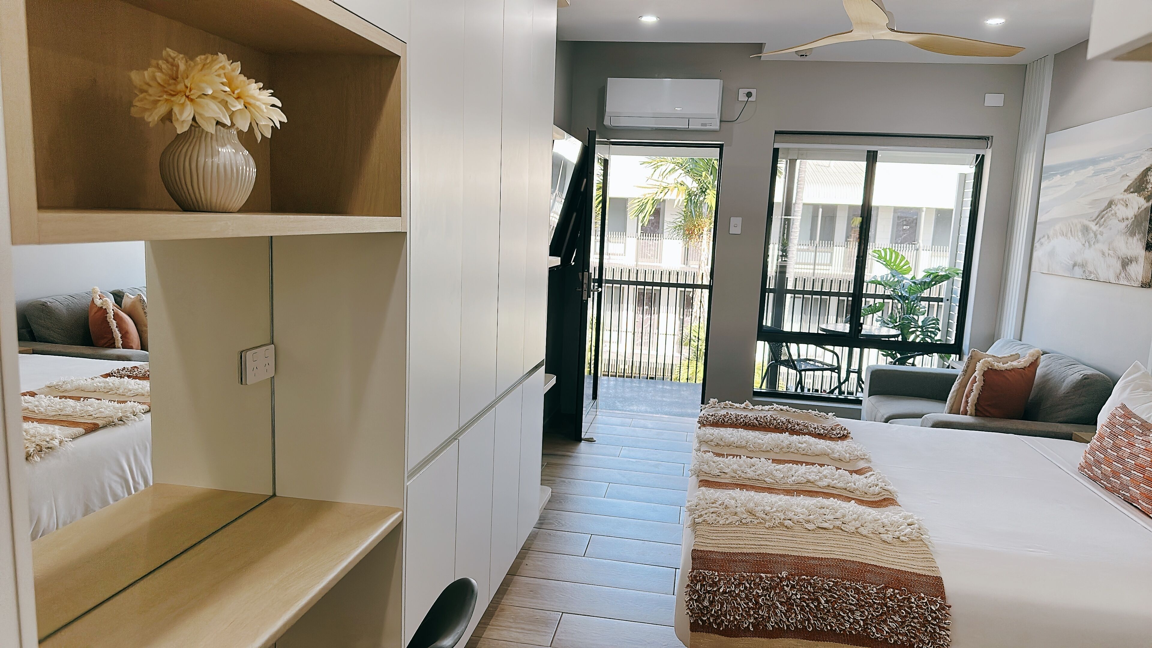 A modern hotel room with a bed, a small desk, a shelf with flowers, a balcony with a view, and an air conditioning unit.