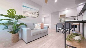 Un salon moderne avec un canapé gris, une grande plante en pot et un coin repas comprenant une table et des chaises.