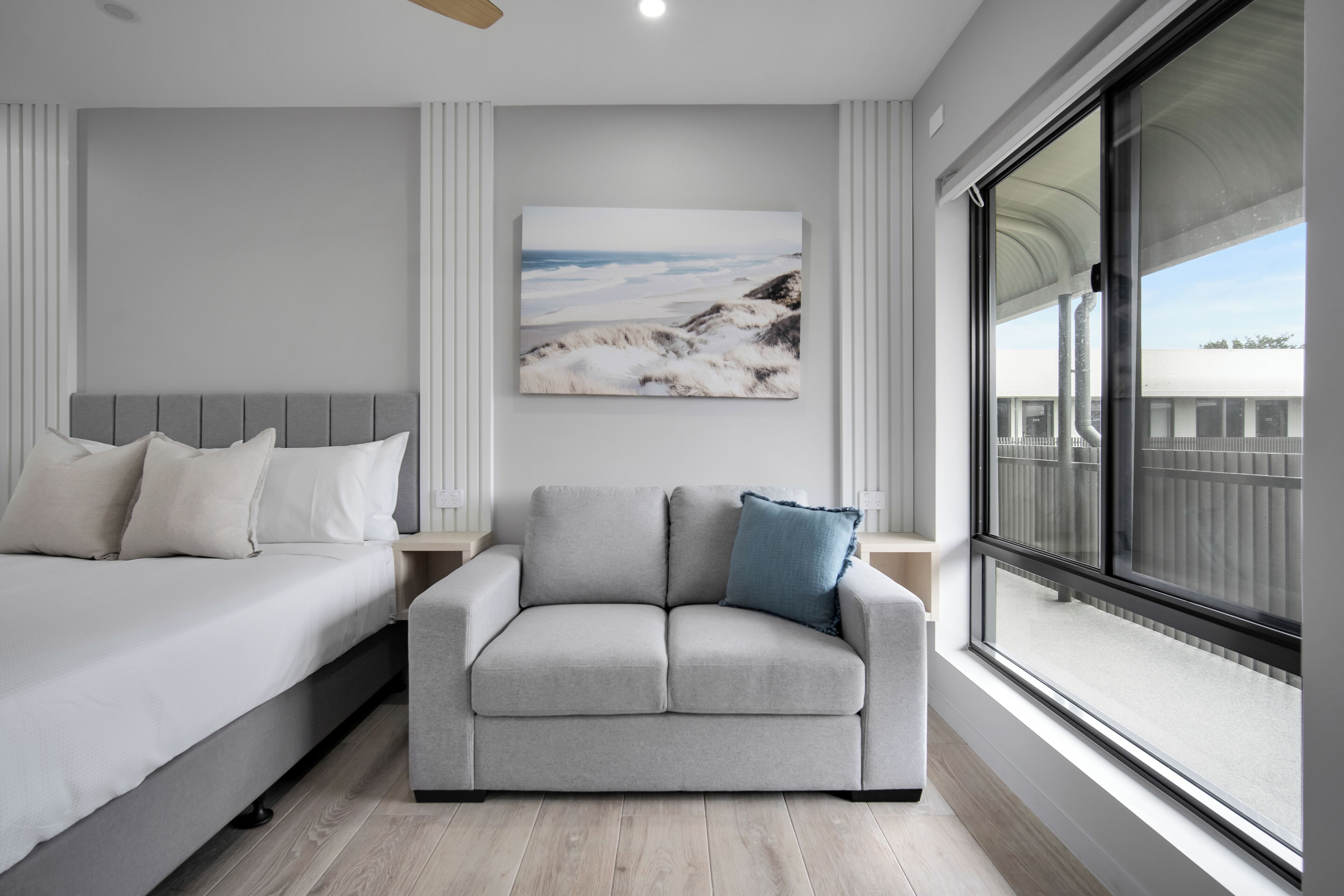 Un dormitorio con una cama, un sofá gris, un ventanal grande y un cuadro en la pared.