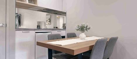 Une cuisine compacte avec un coin repas. Elle comprend une table en bois avec quatre chaises, un vase blanc avec des fleurs et un aménagement de cuisine moderne avec un lave-vaisselle et une bouilloire.