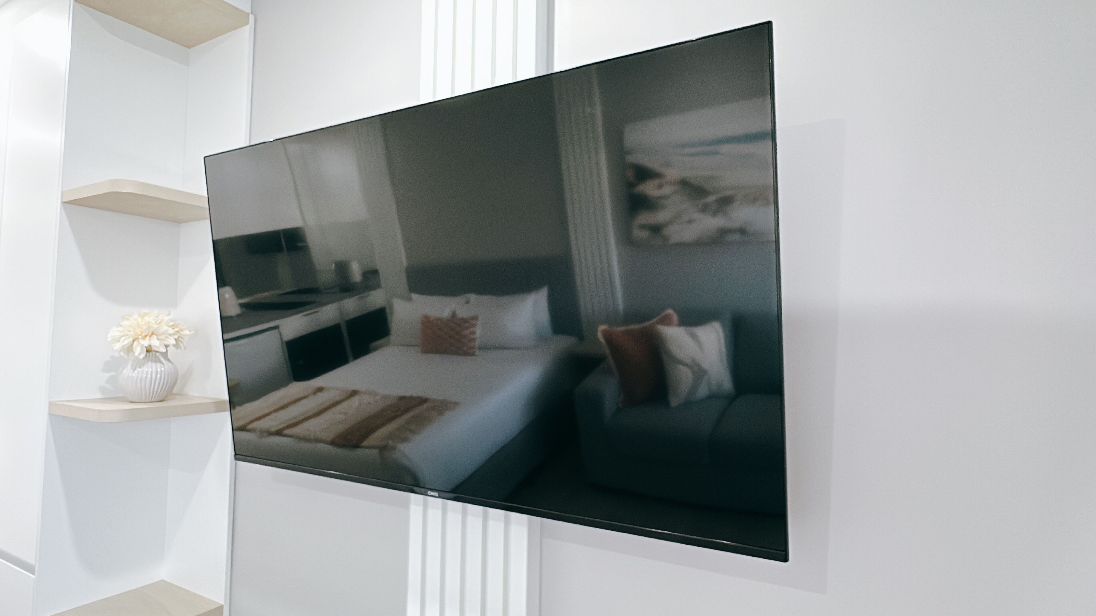 A modern hotel room with a large flat-screen TV mounted on the wall, a bed with a dark headboard, and a small bedside table with a vase of flowers.