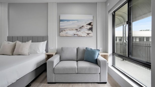 Une chambre moderne avec un lit, un canapé gris et une grande fenêtre donnant sur l’extérieur.