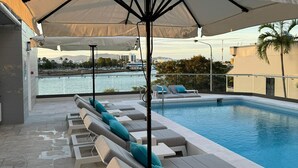 Un espace aménagé au bord de la piscine, avec des chaises longues et des parasols, une piscine, et une vue sur l’eau et les bâtiments situés au loin.