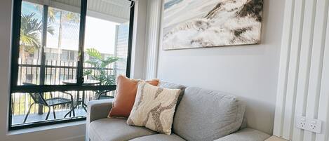 A living room with a grey sofa, a window with blinds, a painting of a beach, and a view of a balcony with plants.