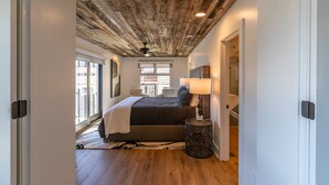 A bedroom with a wooden ceiling, a bed with a gray comforter, a bedside lamp, and a view of the outdoors through a window. - Hawthorn House at Hound Ears - STUNNING Grandfather views! Pool table! Updated! (Boone)