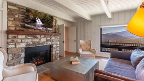 A living room with a stone fireplace, a sofa, a coffee table, and a view of mountains through a large window. - Hawthorn House at Hound Ears - STUNNING Grandfather views! Pool table! Updated! (Boone)