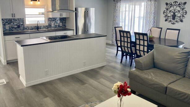 Una cocina moderna con armarios blancos, un refrigerador de acero inoxidable y una encimera negra. Hay un comedor con una mesa y sillas, y un sofá gris en la sala de estar.