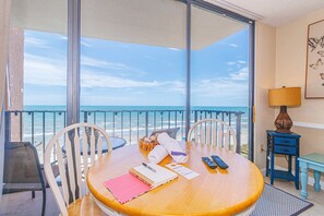 A room with a large window overlooking the beach, a wooden table with a notebook, a lamp, and a remote control.