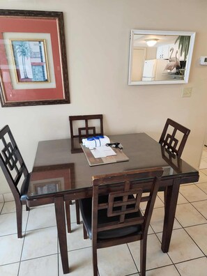 A dining area with a glass-top table, four wooden chairs, a framed picture on the wall, and a kitchen visible in the reflection. - Palace 510 (Myrtle Beach)