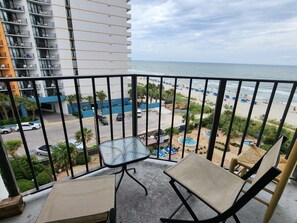 A balcony with a view of a beach, a pool area, and a parking lot.
