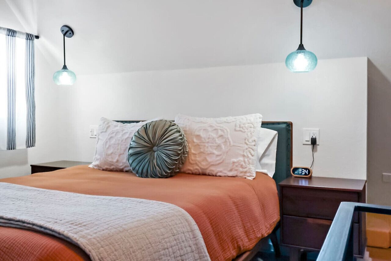 A neatly made bed with an orange bedspread and white pillows, a bedside table with a clock, and two pendant lights.