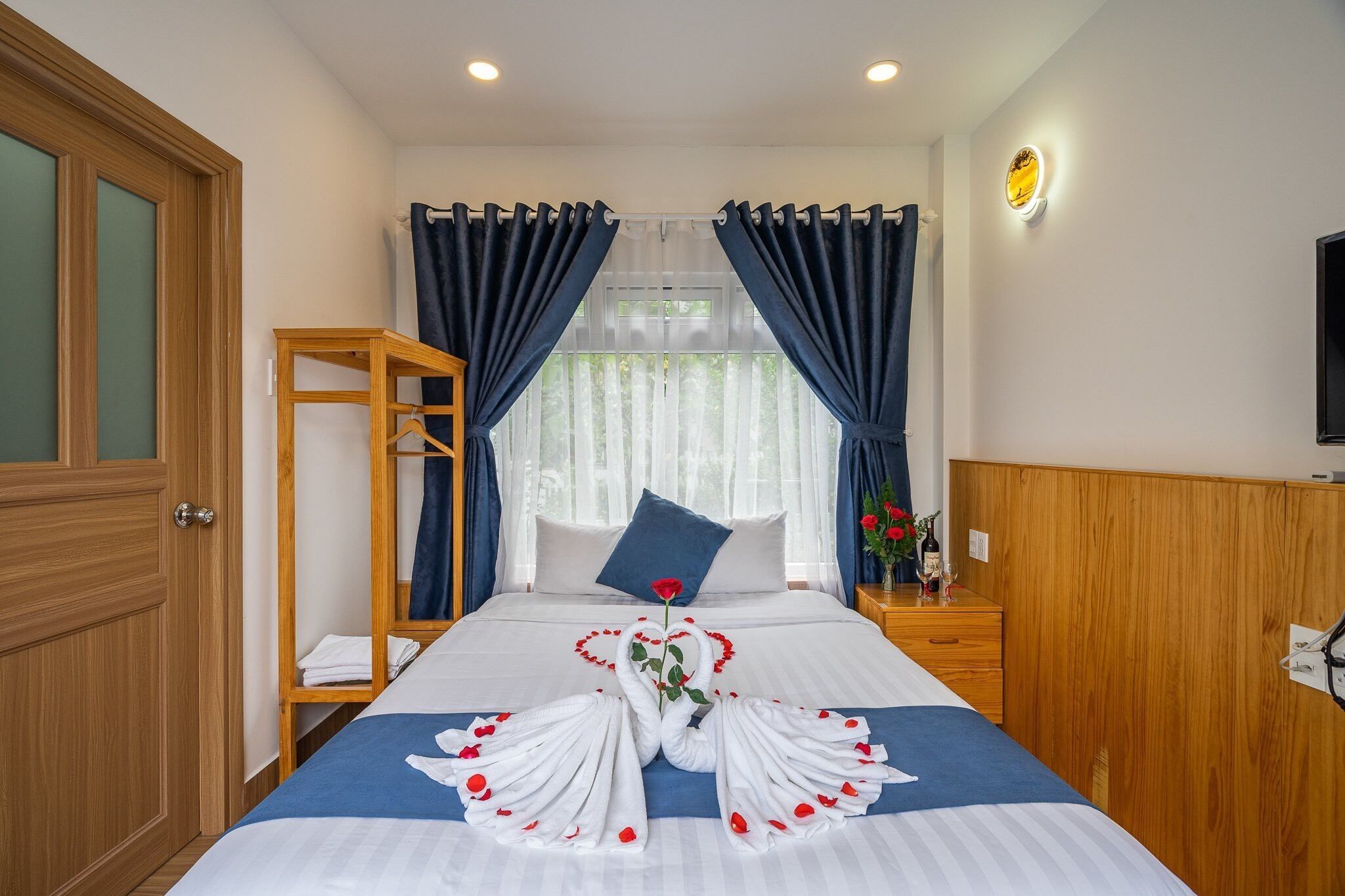 Quarto de hotel com uma cama bem arrumada, uma mesa de cabeceira de madeira com um vaso de flores e um guarda-roupa de madeira.
