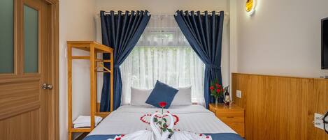 A hotel room with a neatly made bed, a wooden side table with a vase of flowers, and a wooden wardrobe.