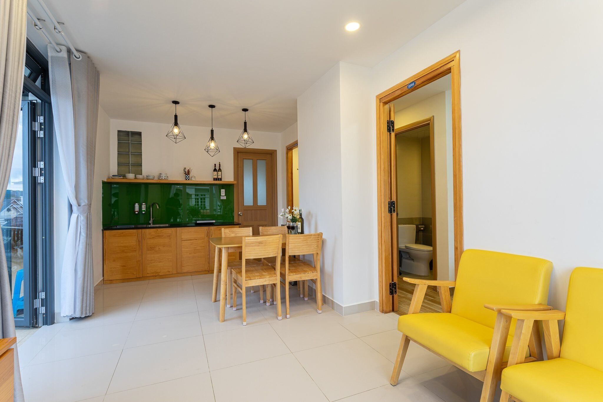 A modern kitchen with wooden cabinets, a dining table, and chairs. There is a yellow sofa in the living area.