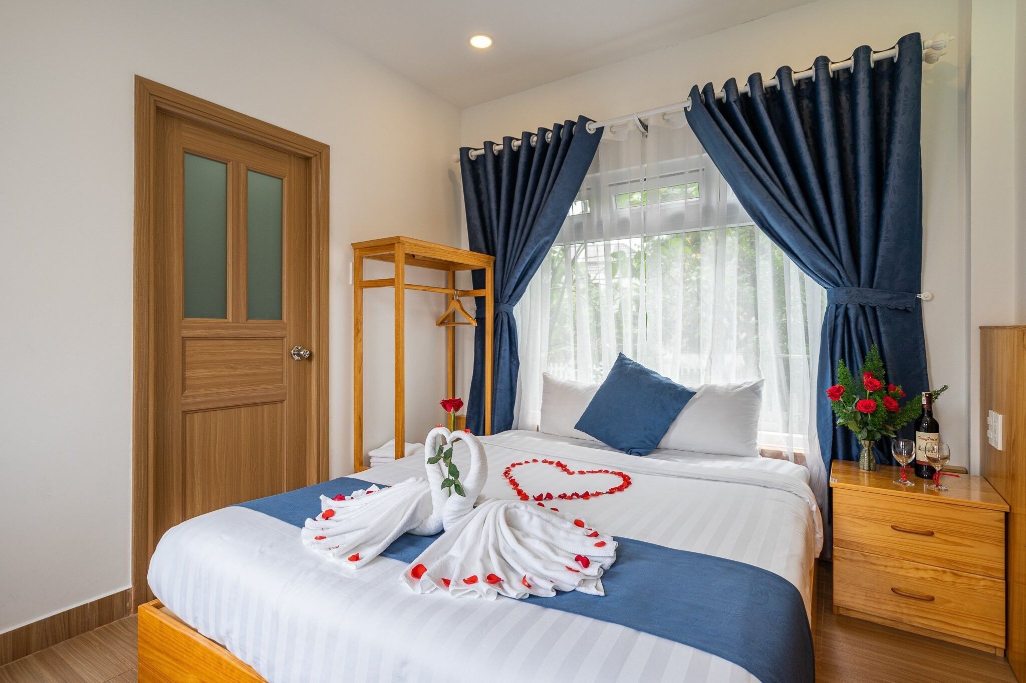 A neatly made bed with a heart-shaped towel arrangement and a swan towel centerpiece, a wooden bedside table with a bottle and glass, and a window with blue curtains.