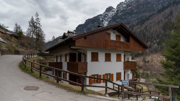 A two-story wooden chalet with balconies, set against a mountainous backdrop.