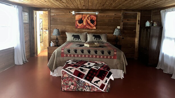 A wooden-paneled bedroom with a bed, two bedside lamps, a wall-mounted security camera, and a large painting. - Beautiful Rustic Cabin 
Just 10 Minutes to Granbury square  (Granbury)