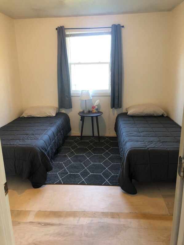 A small room with two beds, a small table with a lamp, and a window with curtains.
