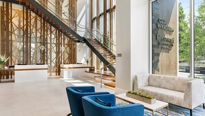 A modern hotel lobby with a reception desk, blue armchairs, a staircase, and a large window with a view of the outdoors.