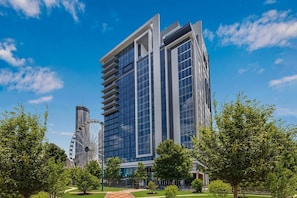 A modern high-rise building with a glass facade, surrounded by trees and a clear blue sky.