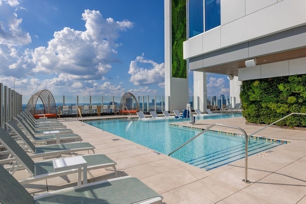 A rooftop pool with lounge chairs, a glass balustrade, and a city skyline view.