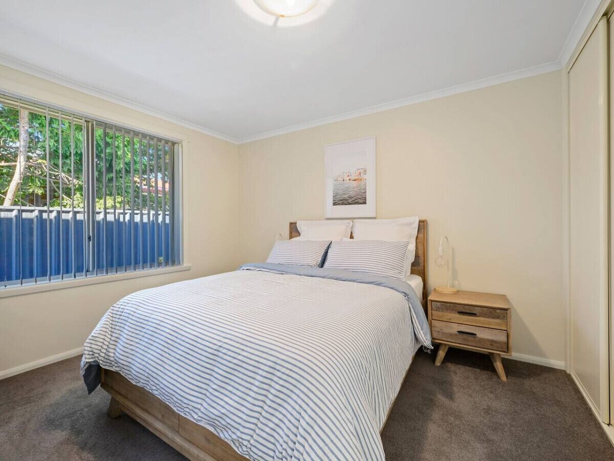 A bedroom with a bed, a nightstand, a window with blinds, and a picture on the wall.