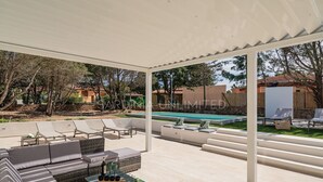 Ein überdachter Außenbereich mit Pool, Liegestühlen und einer Sitzecke mit Kissen und Tisch.