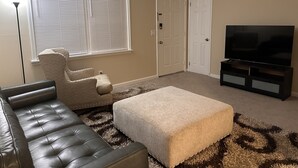 Ein Wohnzimmer mit Ledersofa, Hocker, TV-Schrank und einem großen Fenster mit Jalousien.