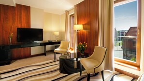 Quarto de hotel com televisão de tela plana, duas cadeiras, uma mesa de centro, um abajur e uma janela grande com vista para edifícios.