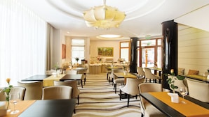 Uma sala de jantar de hotel com várias mesas e cadeiras, um buffet central e uma grande janela com vista para o exterior.