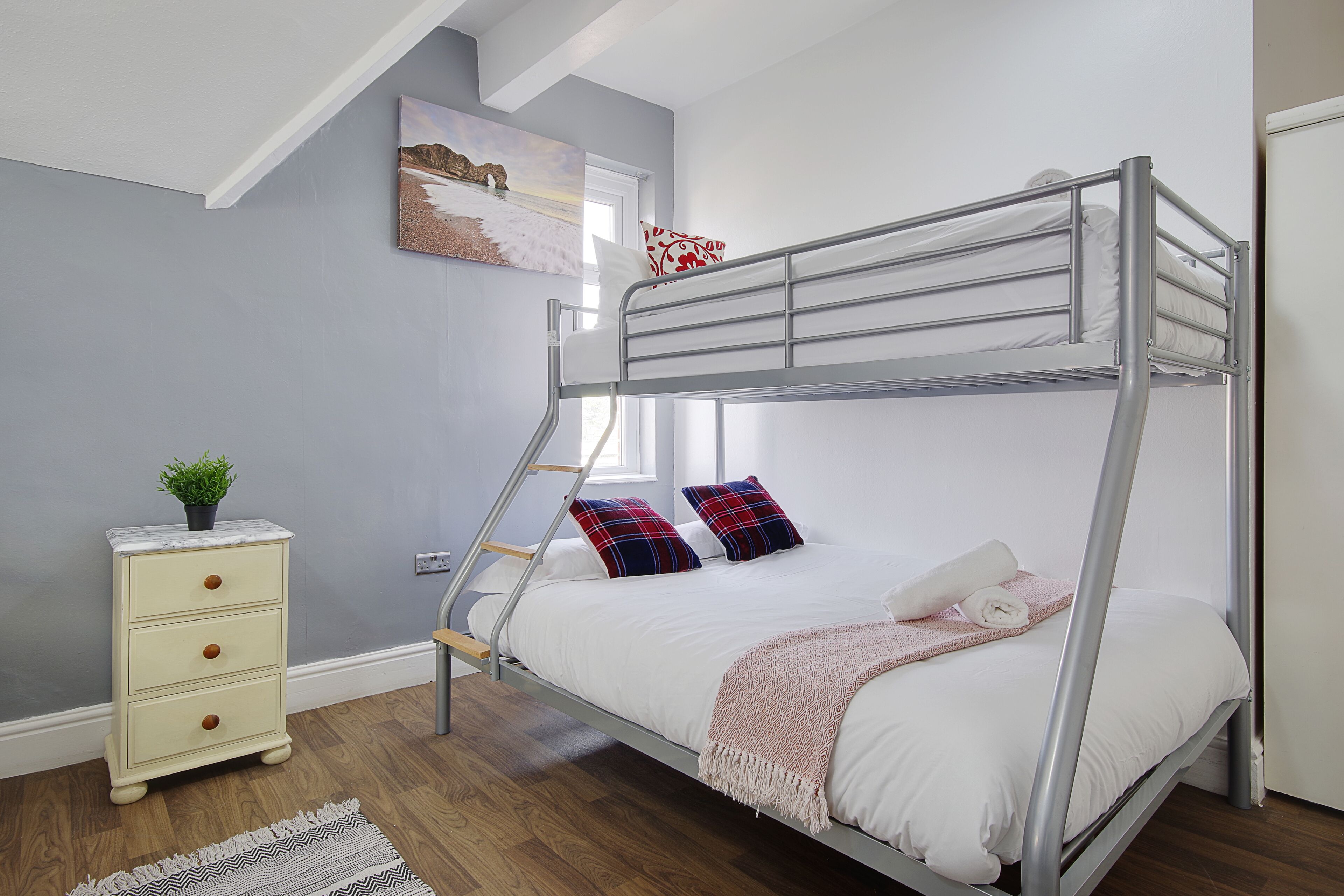 Una cama individual superpuesta con escalera, un aparador, un ventanal y un cuadro en la pared.