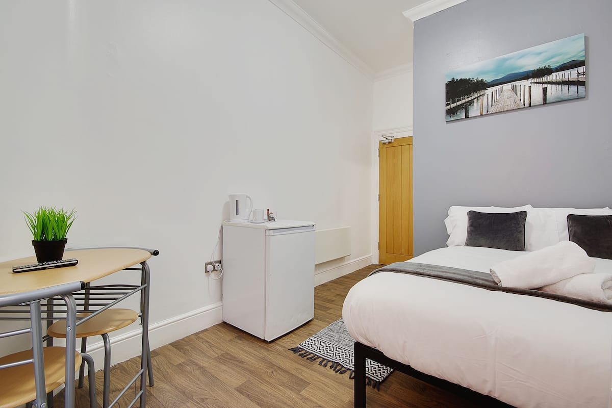 A compact bedroom with a bed, a small desk with a plant, a white refrigerator, and a painting on the wall.