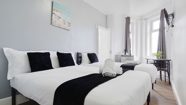 A hotel room with two beds, a desk, a chair, a potted plant, and a window with curtains. - Azure Beach Hotel (Bournemouth)