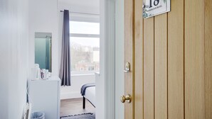 A hotel room with a wooden door, a window with a view of buildings, a bed, a small table with a coffee maker, and a trash bin. - Azure Beach Hotel (Bournemouth)