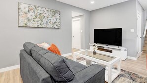 Een woonkamer met een grijze bank, een flatscreen televisie op een witte dressoir en een bloemschilderij aan de muur.