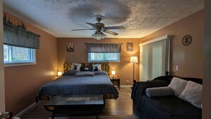 A bedroom with a bed, a bench, a ceiling fan, two bedside tables with lamps, a window, and a door. - Trail Riders Retreat! Hatfield McCoy Trails. Private Pool. Loads of Parking!  (Williamson)