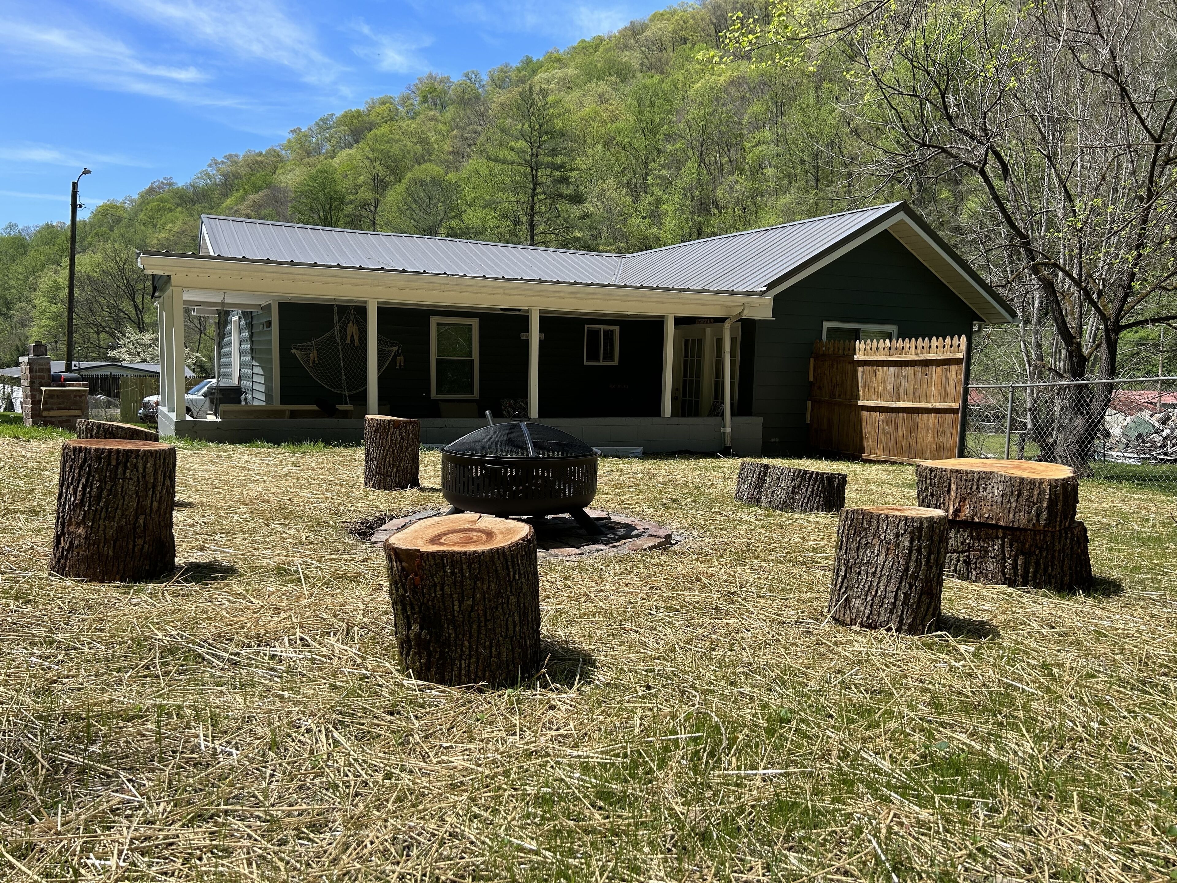 A house with a porch, a fire pit, and several tree stumps in a grassy yard.