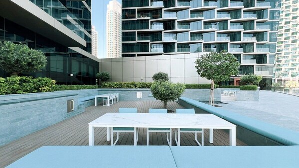 Eine Terrasse im Freien mit einem weißen Tisch und Stühlen, umgeben von Grünflächen und modernen Gebäuden.