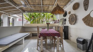 A dining area with a table covered by a patterned tablecloth, wooden chairs, a bench, a ceiling fan, and a view of greenery through the window. - Gajah Villa by Betterplace (Canggu)