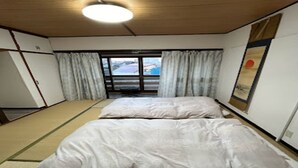 A room with two beds, a tatami mat floor, a sliding door to a balcony, and a ceiling light.