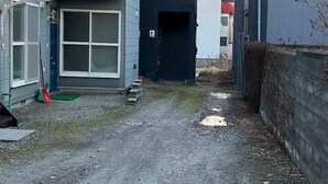 A narrow alleyway between two buildings with a gravel path, a blue door on the left, and a dark-colored building on the right.