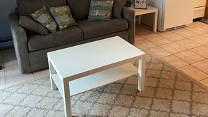 En stue med en grå sofa, et hvidt sofabord og et lille sidebord med en lampe. I baggrunden ses et køkken med træskabe og et hvidt køleskab.