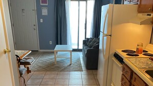 Et kompakt køkken med køleskab, komfur og vask. I det tilstødende opholdsområde er der et spisebord og en sofa.