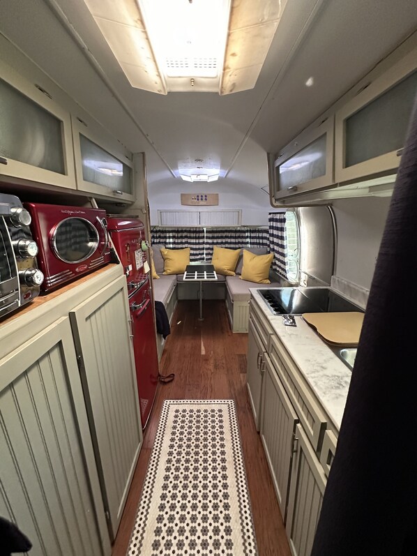 A narrow, compact kitchen with a microwave, stove, and sink. A patterned rug runs along the floor, and the space is illuminated by overhead lights.