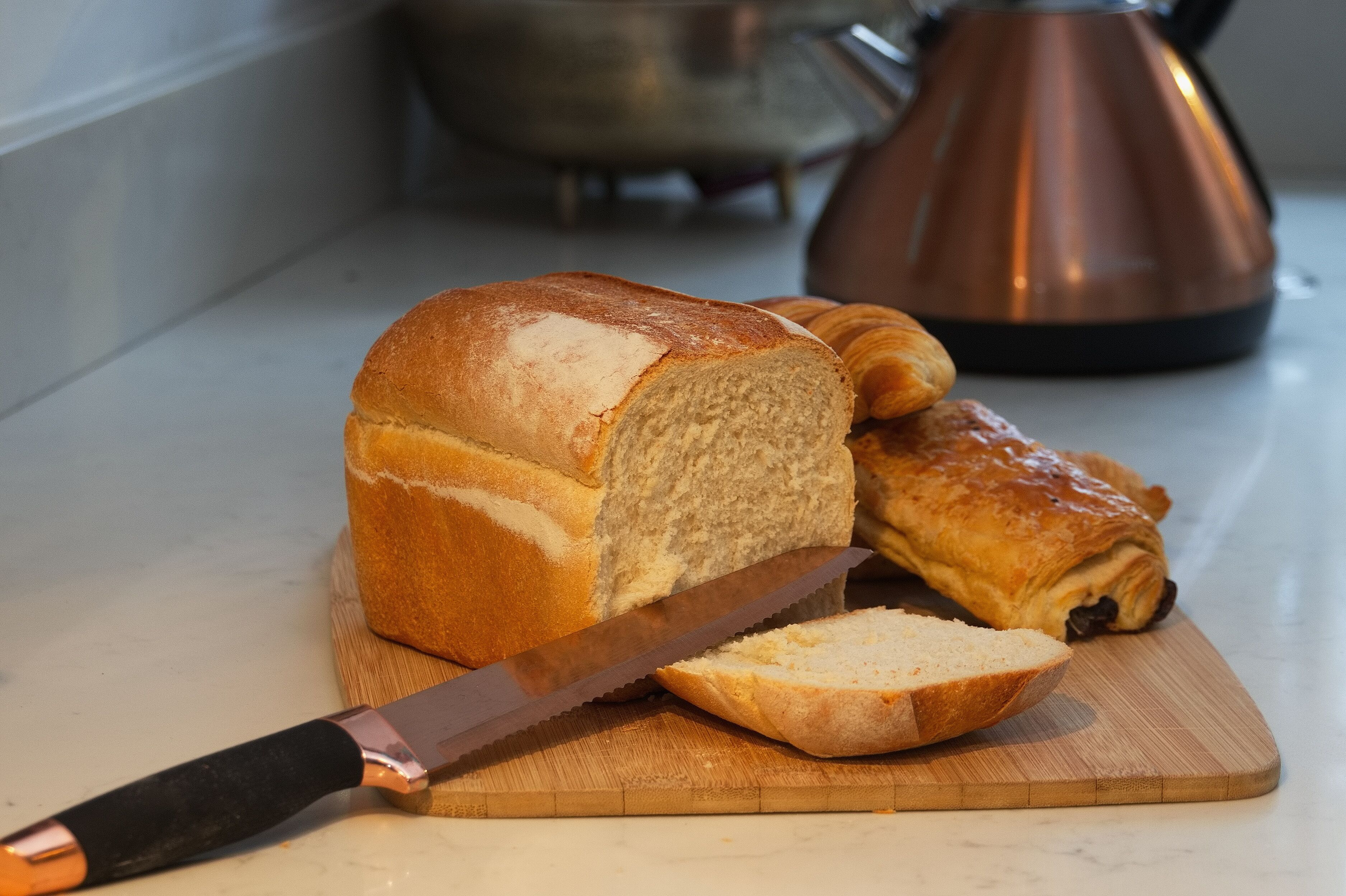 Een brood op een houten snijplank, met een mes, een opgerold gebakje en een koperen pan op de achtergrond.