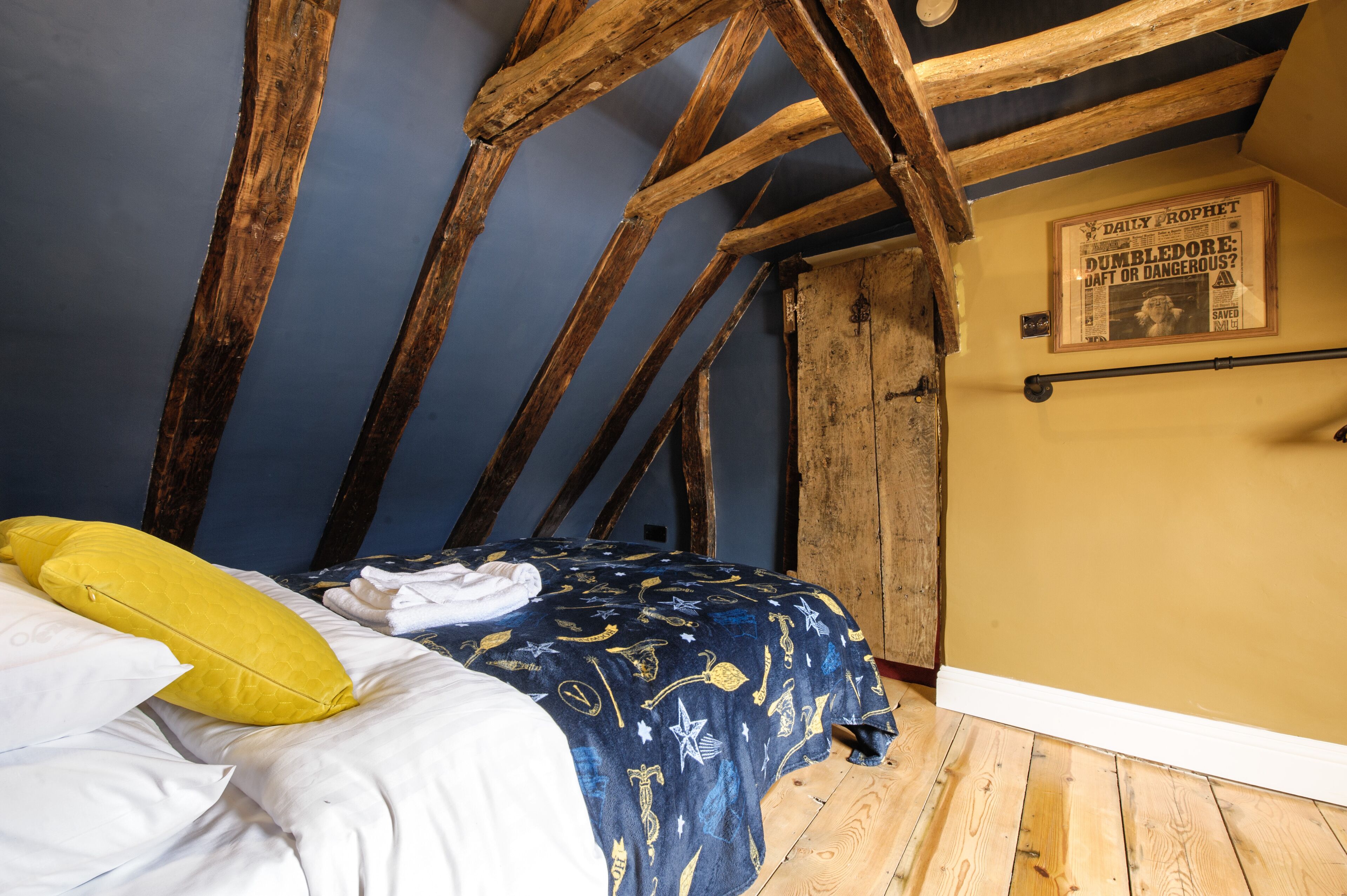 Una habitación con techo inclinado, vigas de madera a la vista, una cama con colcha estampada, un cojín amarillo y un periódico enmarcado en la pared.