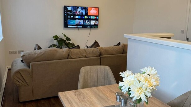 En stue med et træspisebord, beige sofa og et tv på væggen, der viser forskellige kanaler.