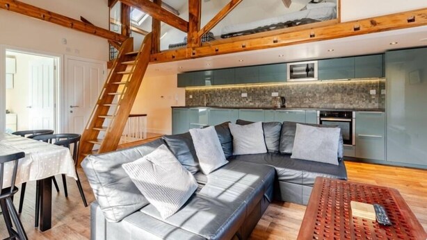 A modern loft-style living space with exposed wooden beams, a built-in kitchen, and a cozy seating area. - Sedbergh Penthouse - KingBed - Tv - Wifi (Sedbergh)