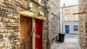 A narrow stone alleyway with a red door and a black trash bin. - Sedbergh Penthouse - KingBed - Tv - Wifi (Sedbergh)